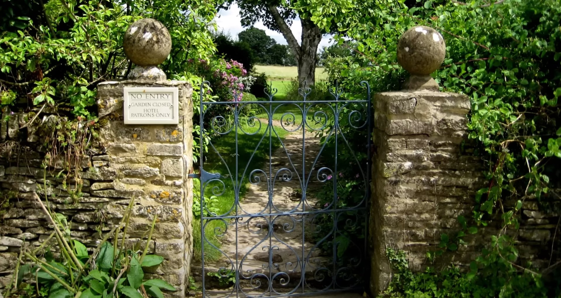 Custom Hardscaping and Landscape Design - Stone Gateway Installation by Maywood Landscaping Elegant stone garden entrance with decorative wrought iron gate, natural stone pillars, and lush green plantings creating an inviting landscape design